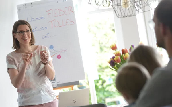 Jana Ahrens steht vor einem Flipchart und erklärt Eltern etwas zum Thema Fieber.