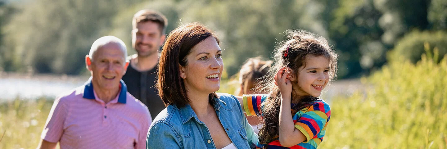 Eine Familie, der drei Generationen angehören, macht einen gemeinsamen Spaziergang. Sie stehen auf einem Feld, im Hintergrund schimmert Wassser, die Sonne scheint. Die Mutter hält ihre Tochter fest dem Arm. Beide lachen.