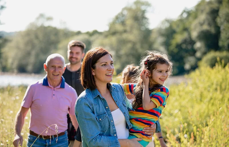 Eine Familie, der drei Generationen angehören, macht einen gemeinsamen Spaziergang. Sie stehen auf einem Feld, im Hintergrund schimmert Wassser, die Sonne scheint. Die Mutter hält ihre Tochter fest dem Arm. Beide lachen.