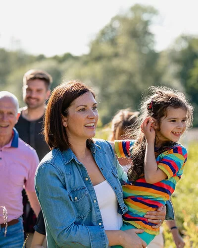 Eine Familie, der drei Generationen angehören, macht einen gemeinsamen Spaziergang. Sie stehen auf einem Feld, im Hintergrund schimmert Wassser, die Sonne scheint. Die Mutter hält ihre Tochter fest dem Arm. Beide lachen.