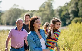 Eine Familie, der drei Generationen angehören, macht einen gemeinsamen Spaziergang. Sie stehen auf einem Feld, im Hintergrund schimmert Wassser, die Sonne scheint. Die Mutter hält ihre Tochter fest dem Arm. Beide lachen.