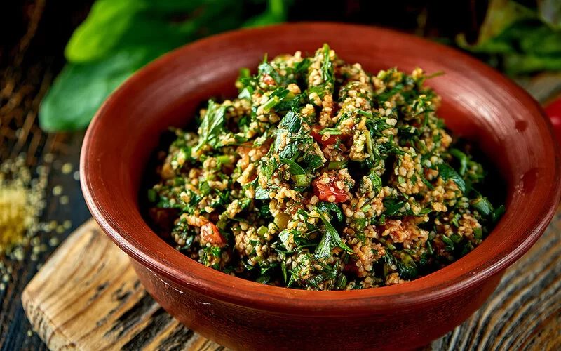 In einer runden Schüssel ist Tabouleh angerichtet. Der Salat enthält Bulgur, frische Minzblätter, gehackte Petersilie, gestückelte Tomaten und Zwiebeln.