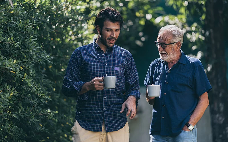 Ein Vater geht mit seinem erwachsenen Sohn spazieren, sie sind in ein freundliches Gespräch vertieft. Der Sohn hält eine Tasse in der Hand. 