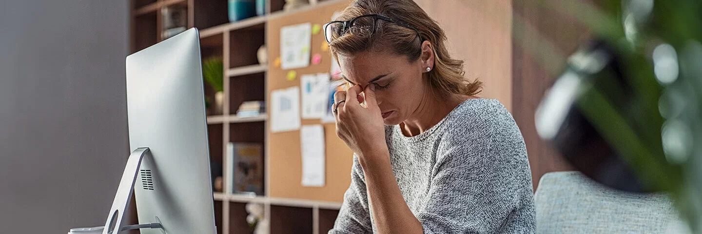 Eine Frau mit einem grauen Pullover bekleidet sitzt an einem Schreibtisch. Sie stützt den Ellbogen auf die Tischfläche und hält sich den Nasenrücken.