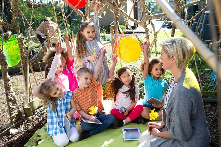 Eine Lehrerin mit rot-blonden Haaren, blauer Bluse und einem Tablet in den Händen lächelt in die Kamera. Im Hintergrund ist ein Klassenzimmer mit Kindern zu sehen, die an ihren Plätzen arbeiten.