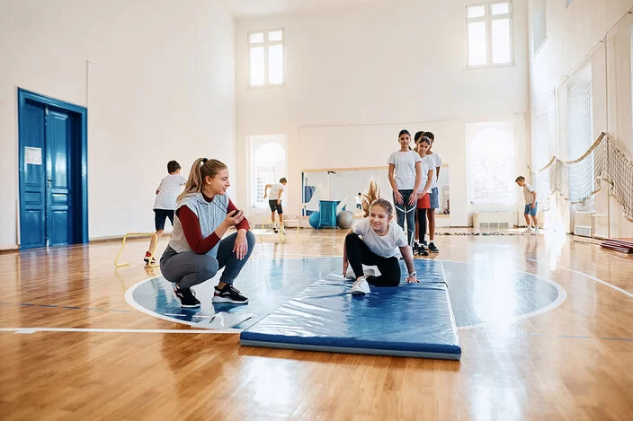 Kinder turnen in einer großen, hellen Turnhalle. Im Vordergrund ist ein Mädchen auf einer blauen Matte zu sehen. Neben ihr hockt die Sportlehrerin mit einer Stoppuhr in der Hand. Hinter der Matte warten andere Kinder in einer Reihe.