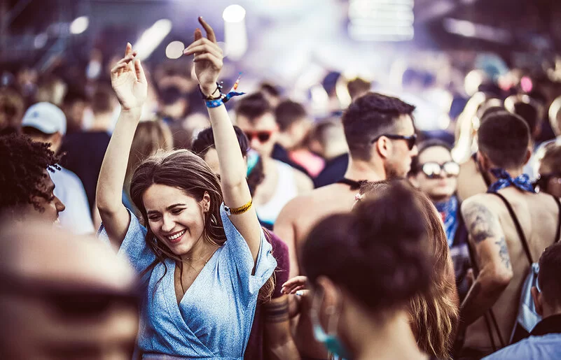 Eine junge Frau in einem blauen Sommerkleid tanzt, die Hände in die Höhe gereckt und lächelnd, in einer Menschenmenge.