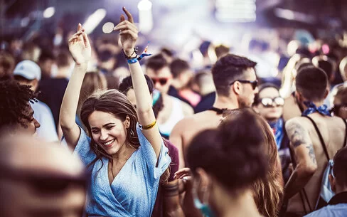 Eine junge Frau in einem blauen Sommerkleid tanzt, die Hände in die Höhe gereckt und lächelnd, in einer Menschenmenge.