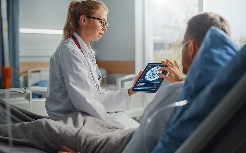 Eine junge Ärztin sitzt auf der Bettkante eines Patienten, der sich nach einer Operation erholt. Sie hält ein Tablet in der Hand, auf dem ein Gehirnscan zu sehen ist und erklärt das Ergebnis. Der Mann tippt mit Daumen und Zeigefinder der rechten Hand auf den Bildschirm.