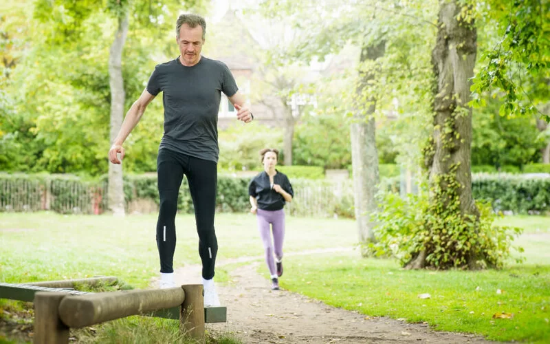 Ein Mann in dunkler Sportkleidung steht auf einem Balken im Park. Hinter ihm joggt eine Frau in Sportkleidung.