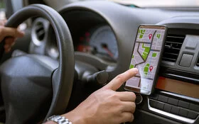 Ein Mann lenkt mit einer Hand ein Auto und bedient mit der anderen Hand die Navigation auf einem Handy.