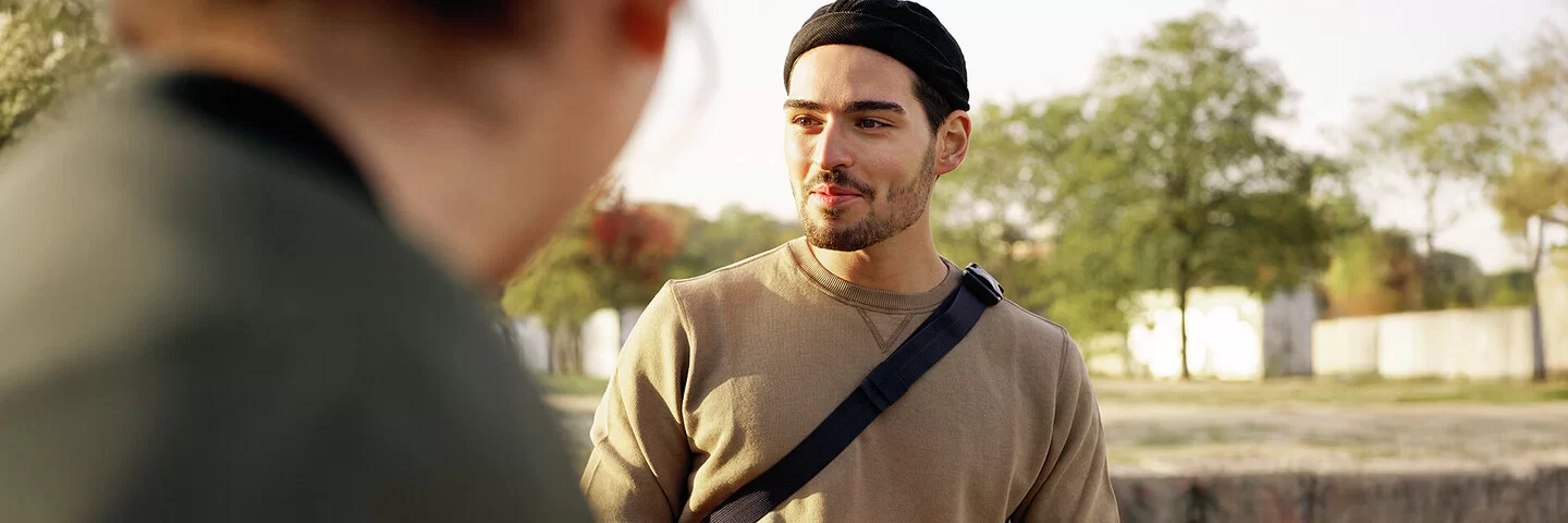 Ein junger Mann ist im Park unterwegs und unterhält sich mit einer anderen Person. In der Hand hält er ein Smartphone.