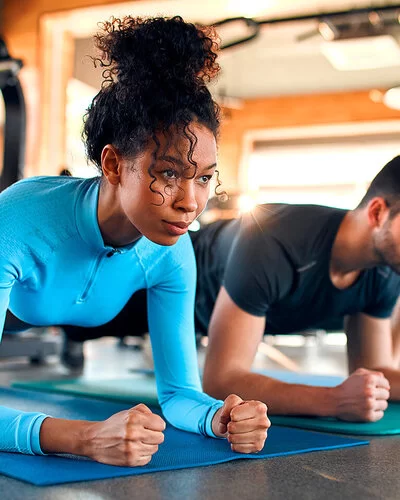 Drei Personen, eine Frau, ein Mann und eine Frau, machen Plank-Übungen auf Sportmatten in einem Fitnessstudio.