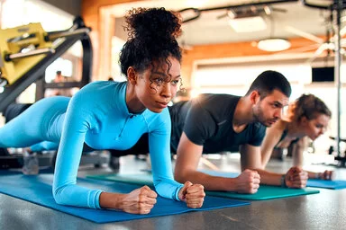 Drei Personen, eine Frau, ein Mann und eine Frau, machen Plank-Übungen auf Sportmatten in einem Fitnessstudio.