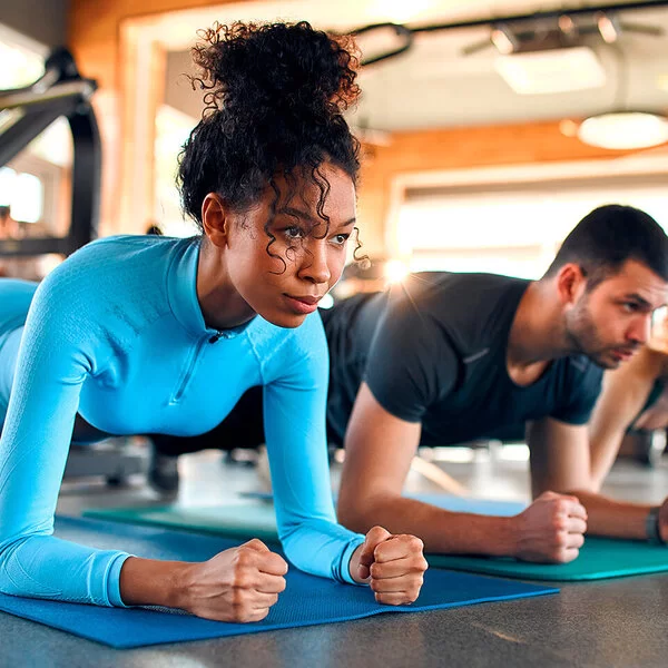 Drei Personen, eine Frau, ein Mann und eine Frau, machen Plank-Übungen auf Sportmatten in einem Fitnessstudio.