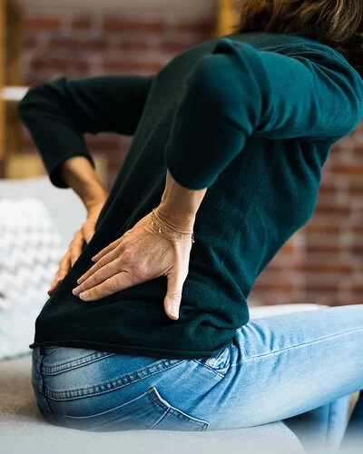 Eine Frau sitzt vorgebeugt auf einer Couch und hält sich mit beiden Händen den unteren Rücken.