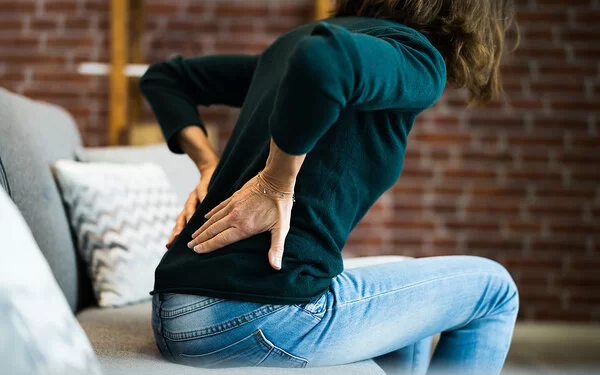 Eine Frau sitzt vorgebeugt auf einer Couch und hält sich mit beiden Händen den unteren Rücken.