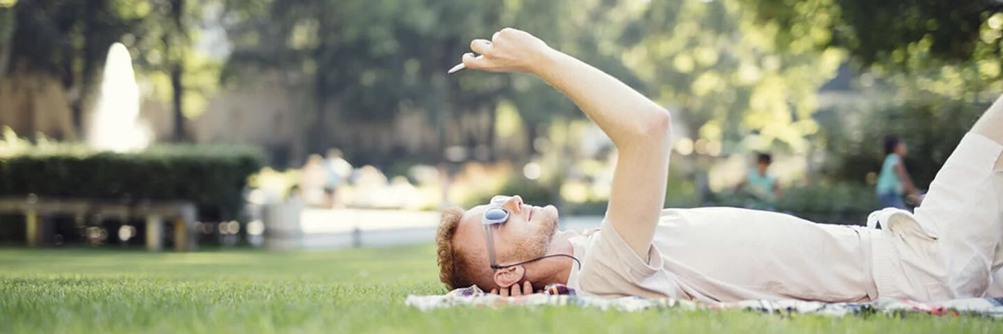 Ein junger Mann liegt auf einer Decke im Gras und blickt auf sein Mobiltelefon.