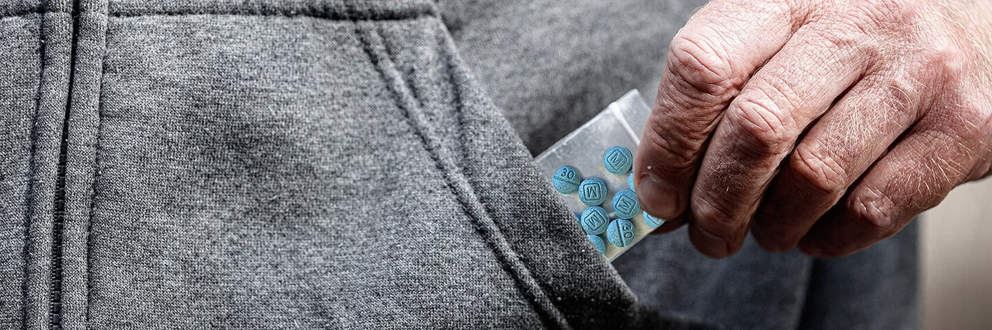 Blaue Pillen in einer kleinen Plastiktüte, die von einer Männerhand in Nahaufnahme aus einer Jackentasche gezogen wird.