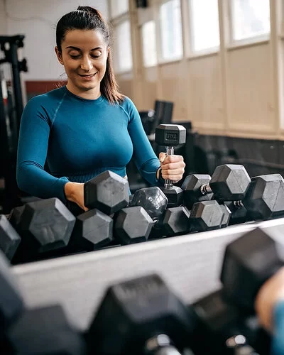 Eine junge Frau in Trainingskleidung steht in einem Fitnessstudio vor einem Regal mit zahlreichen Hanteln und sucht sich lächelnd ein Paar zum Trainieren aus.