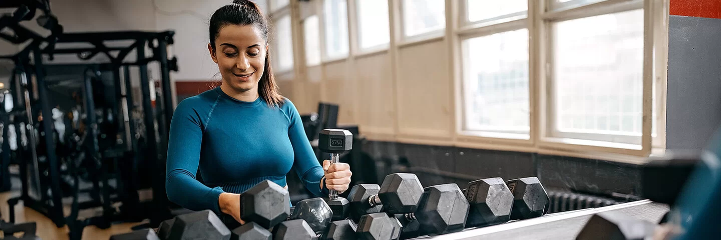 Eine junge Frau in Trainingskleidung steht in einem Fitnessstudio vor einem Regal mit zahlreichen Hanteln und sucht sich lächelnd ein Paar zum Trainieren aus.