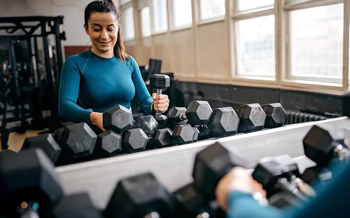 Eine junge Frau in Trainingskleidung steht in einem Fitnessstudio vor einem Regal mit zahlreichen Hanteln und sucht sich lächelnd ein Paar zum Trainieren aus.