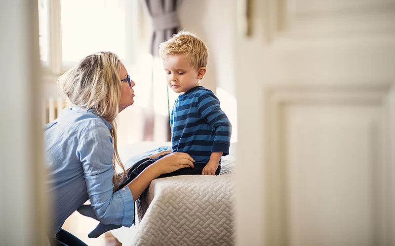 Eine Frau mit langend blonden Haaren und Brille hockt vor einem Bett und spricht mit einem kleinen blonden Jungen in einem blaugestreifeten Pullover, der auf der Bettkante sitzt.