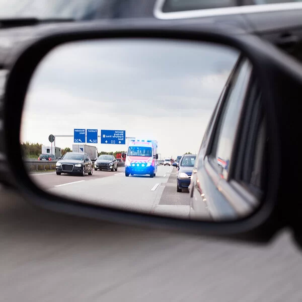 Nahaufnahme eines Auto-Seitenspiegels. Darin ist ein Krankenwagen, der von hinten näherkommt. Die Autos auf der Autobahn haben eine ordnungsgemäße Rettungsgasse gebildet.