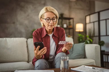 Eine blonde Frau mit Brille und bequemer Alltagskleidung sitzt auf einem Sofa, sie ist etwa Ende 50. Im Hintergrund ist eine moderne Einrichtung zu erkennen. Die Frau hält in ihrer linken Hand eine Blisterverpackung mit Tabletten, in ihrer rechten Hand ein Smartphone. Die Frau blickt prüfen auf die Rückseite der Blisterverpackung.