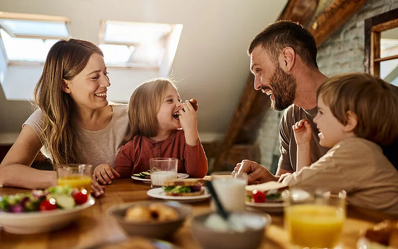 Eine Familie mit zwei Kindern sitzt entspannt und lachend zusammen beim Essen an einem Holztisch.