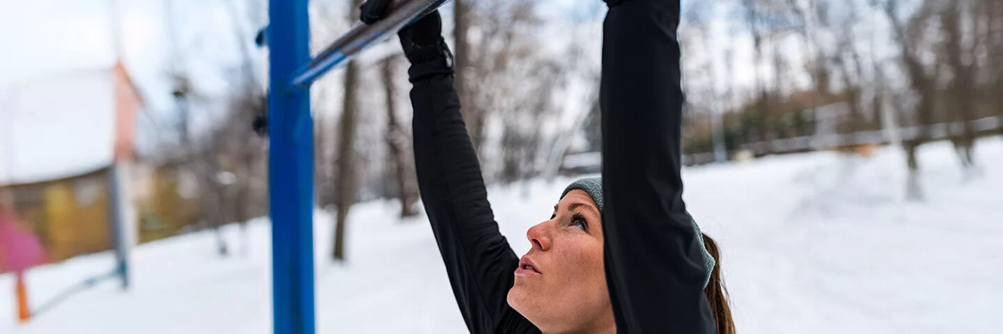 Nahaufnahme einer Frau, die sich im Winter an einer Klimmzugstange in einem Park festhält.