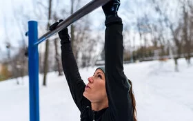 Nahaufnahme einer Frau, die sich im Winter an einer Klimmzugstange in einem Park festhält.