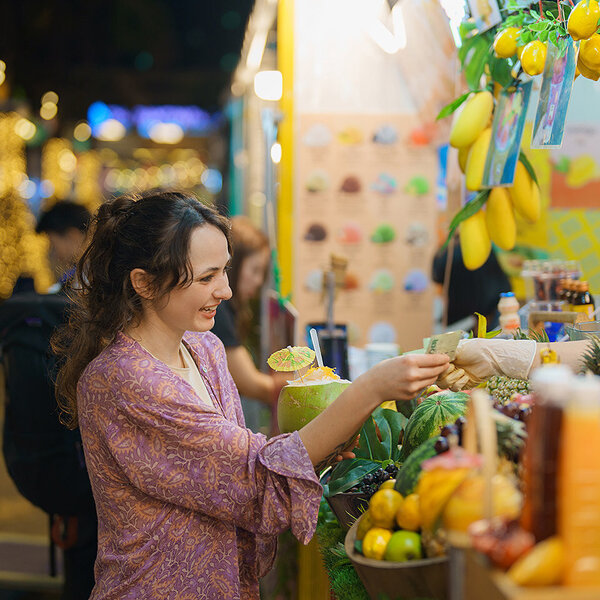 Eine junge Frau auf einem Markt in einem tropischen Land kauft ein Getränk in einer Kokosnuss, dekoriert mit einem Schirmchen.