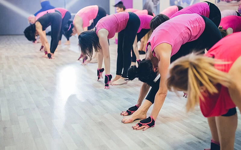 Teilnehmerinnen einer Piloxing-Gruppe beugen sich nach vorne und berühren mit den Händen den Hallenboden. Sie tragen fingerlose, schwarz-pinke Handschuhe und Sportkleidung in Schwarz und Pink.