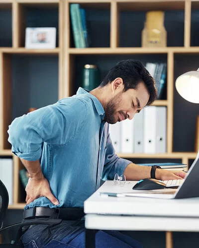 Ein junger Mann sitzt am Schreibtisch in seinem Arbeitszimmer oder Büro. Er hat die Augen geschlossen und fasst sich mit der rechten Hand in die Lendengegend.