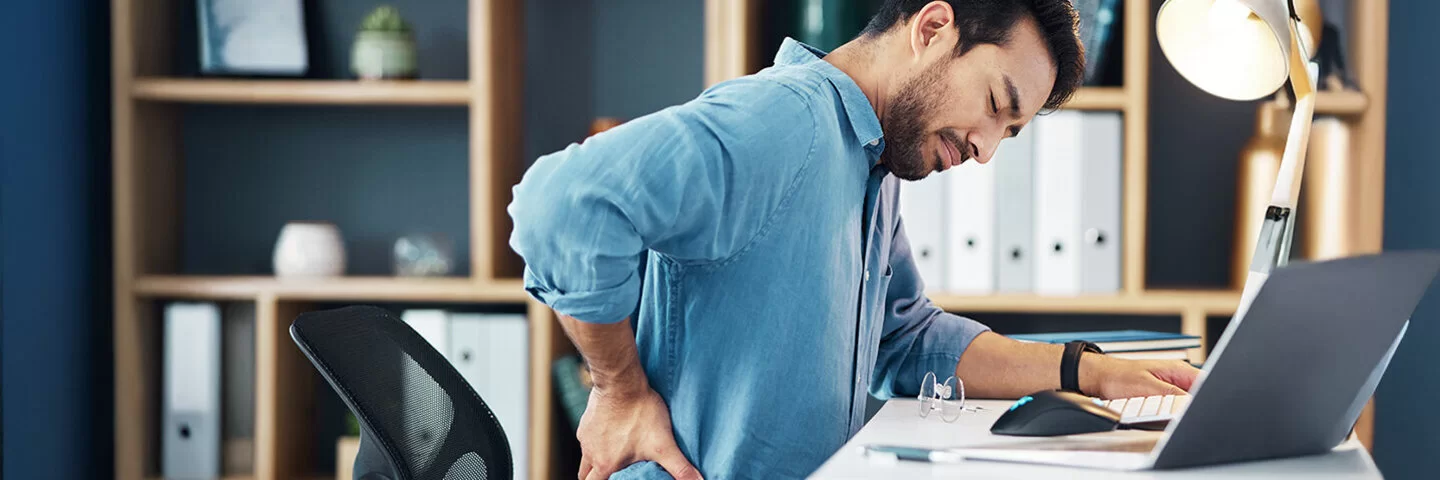 Ein junger Mann sitzt am Schreibtisch in seinem Arbeitszimmer oder Büro. Er hat die Augen geschlossen und fasst sich mit der rechten Hand in die Lendengegend.