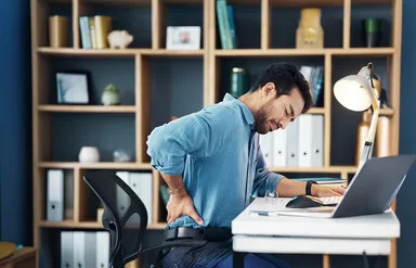 Ein junger Mann sitzt am Schreibtisch in seinem Arbeitszimmer oder Büro. Er hat die Augen geschlossen und fasst sich mit der rechten Hand in die Lendengegend.