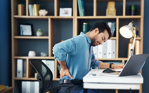 Ein junger Mann sitzt am Schreibtisch in seinem Arbeitszimmer oder Büro. Er hat die Augen geschlossen und fasst sich mit der rechten Hand in die Lendengegend.