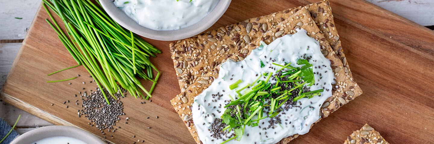 Auf Holzbrettchen liegen Scheiben körnigen Knäckebrots mit frischem Quark und Kräutern.