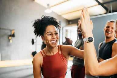 Eine junge Frau in Sportbekleidung gibt einer anderen Frau ein High Five in einem Fitnessstudio. Im Hintergrund stehen zwei fröhlich lachende Männer.