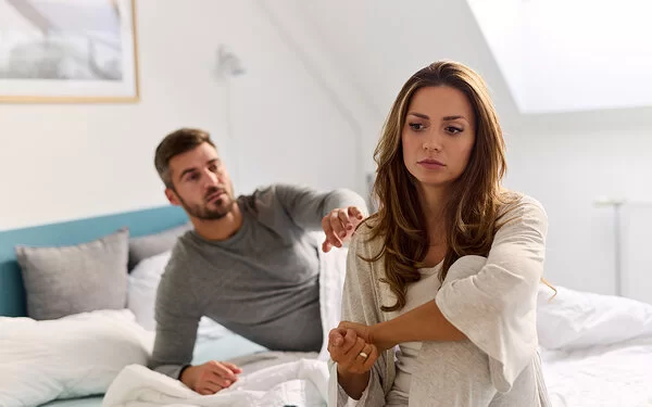 Ein Mann liegt mit dem rechten Arm aufgestützt im Bett und berührt mit der linken Hand die Schulter einer Frau, die auf der Bettkante sitzt und betrübt schaut.