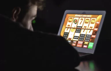 Eine nicht erkennbare Person sitzt im Dunkeln vor einem Notebook und schaut auf ein Glücksspiel eines Online-Casinos.