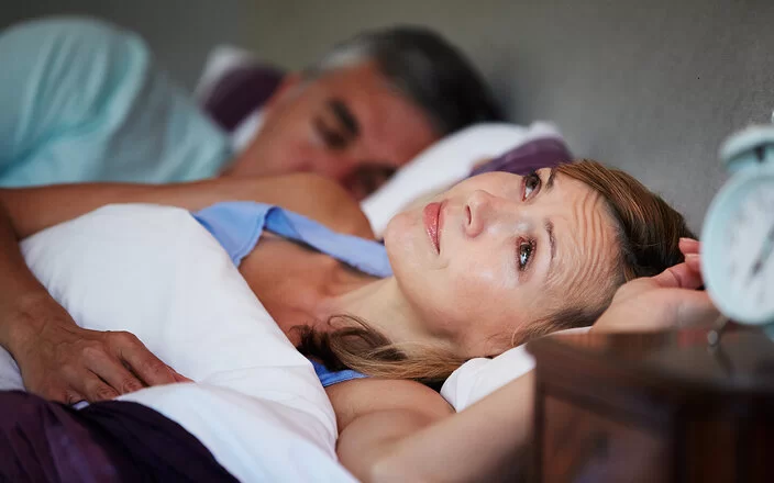 Ein Schlafzimmer bei Nacht. Mann und Frau liegen im Bett. Der Mann schläft und die Frau liegt wach mit Sorgenfalten auf der Stirn.