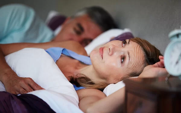 Ein Schlafzimmer bei Nacht. Mann und Frau liegen im Bett. Der Mann schläft und die Frau liegt wach mit Sorgenfalten auf der Stirn.