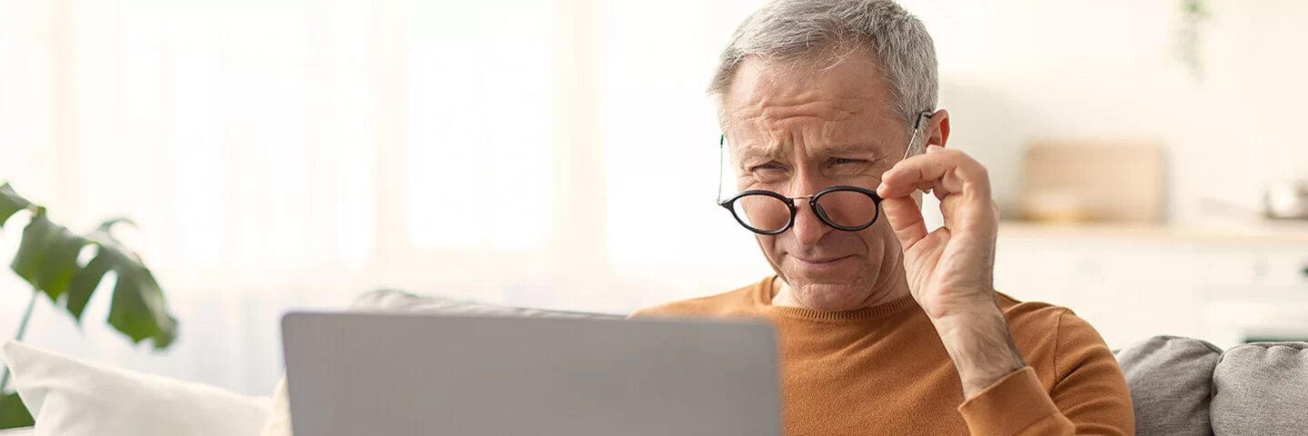 Ein älterer Mann sitzt auf einem Sofa mit einem Laptop auf dem Schoß. Über den Rand seiner nach unten gezogenen Brille blickt er mit angestrengt zusammengekniffenen Augen auf den Bildschirm.