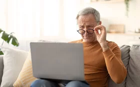 Ein älterer Mann sitzt auf einem Sofa mit einem Laptop auf dem Schoß. Über den Rand seiner nach unten gezogenen Brille blickt er mit angestrengt zusammengekniffenen Augen auf den Bildschirm.