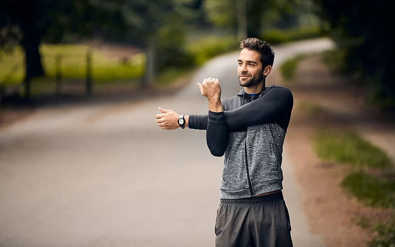 Ein junger Mann steht in Trainingskleidung auf einer Straße, die durch einen Wald führt. Lächelnd macht er Dehnübungen mit den Armen.