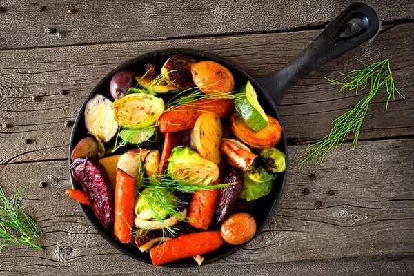 Auf einem rustikalen Holztisch steht eine gusseiserne Pfanne mit Möhren, Kartoffeln, Zwiebeln, Zucchini, roter Beete und anderem gesunden Herbstgemüse.