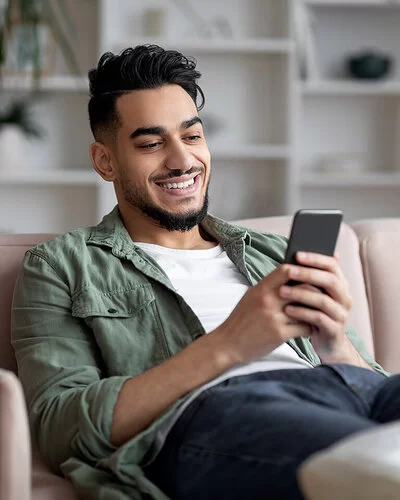 Ein junger Mann sitzt gemütlich zuhause auf dem Sofa und spielt auf dem Smartphone. Er lacht, es macht ihm Spaß.