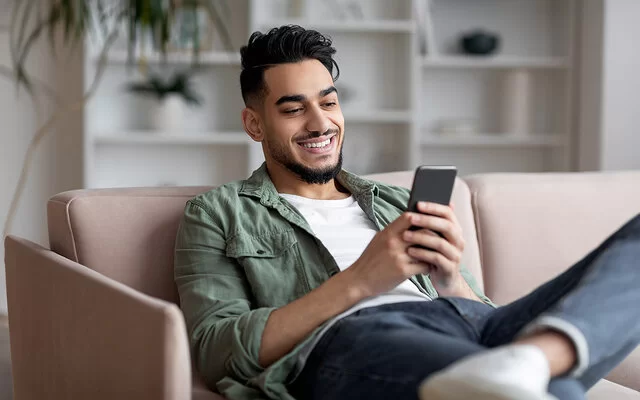 Ein junger Mann sitzt gemütlich zuhause auf dem Sofa und spielt auf dem Smartphone. Er lacht, es macht ihm Spaß.